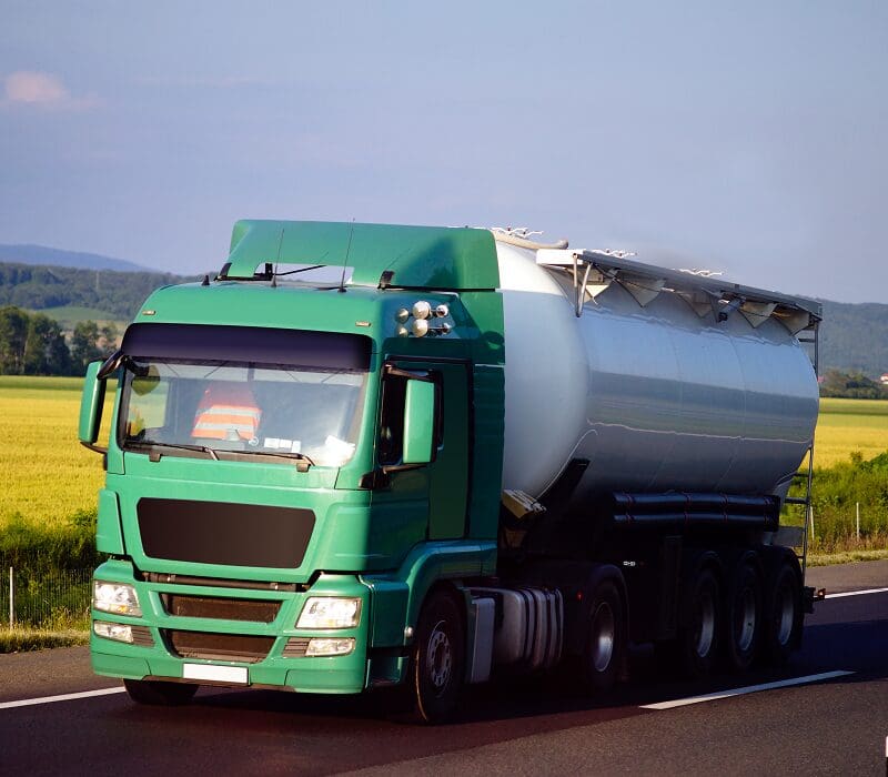 Green and white tanker truck driving down a road surrounded by a field of crops, trees